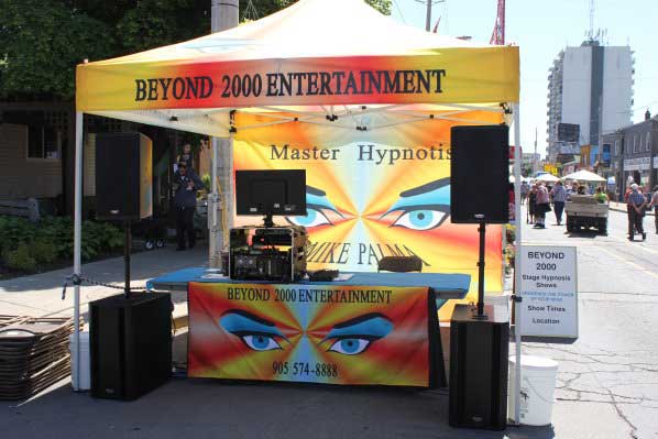 Hypnosis Hamilton, Hamilton DJ, Beyond 2000 Entertainments Canopy with back wall. Very colorful canopy with hypnotic eyes on all four side of the roof and Beyond 2000 Entertainment written on the canopy edges. Back drop has large hypnotic eyes. With the words Master hypnotist above the eyes. Below the eyes Mike Palma Experience the power of your mind. Table set up with sound equipment & computer on it. Table skirting has hypnotic eyes, Beyond 2000 Entertainment and business phone number (905) 574-8888. Taken in Hamilton Ontario